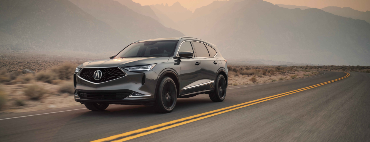 3/4 view of a grey MDX driving down a road in a mountainous desert landscape. / Vue 3/4 d’un MDX gris circulant sur une route dans un paysage montagneux désert.