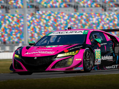 Close-up of a pink, sponsorship decaled Acura NSX GT3 on a racetrack with colour-dotted stadium seating in the background. Gros plan d’une Acura NSX GT3 rose ornée de décalcomanies de commanditaires roulant sur un circuit de course avec des estrades multicolores en arrière-plan.