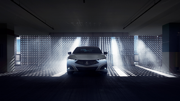 Front view of a white Acura TLX in a darkened garage with natural light shining through a gated wall behind. // Vue avant d’une Acura TLX blanche dans un garage sombre devant un mur qui laisse passer la lumière naturelle.
